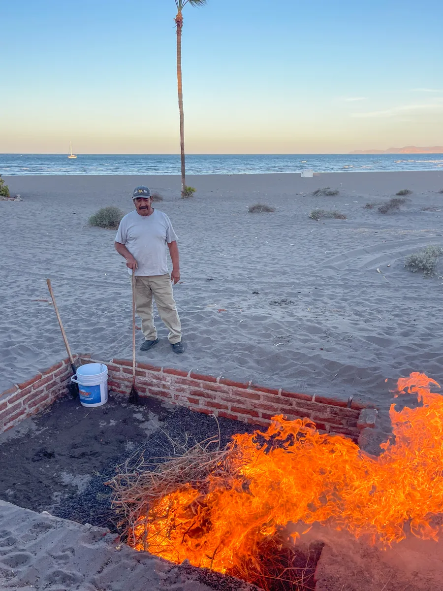 man standing next to a brick pit in the sand at the beach with a large fire in the middle of the pit and the sun setting over the horizon behind him.