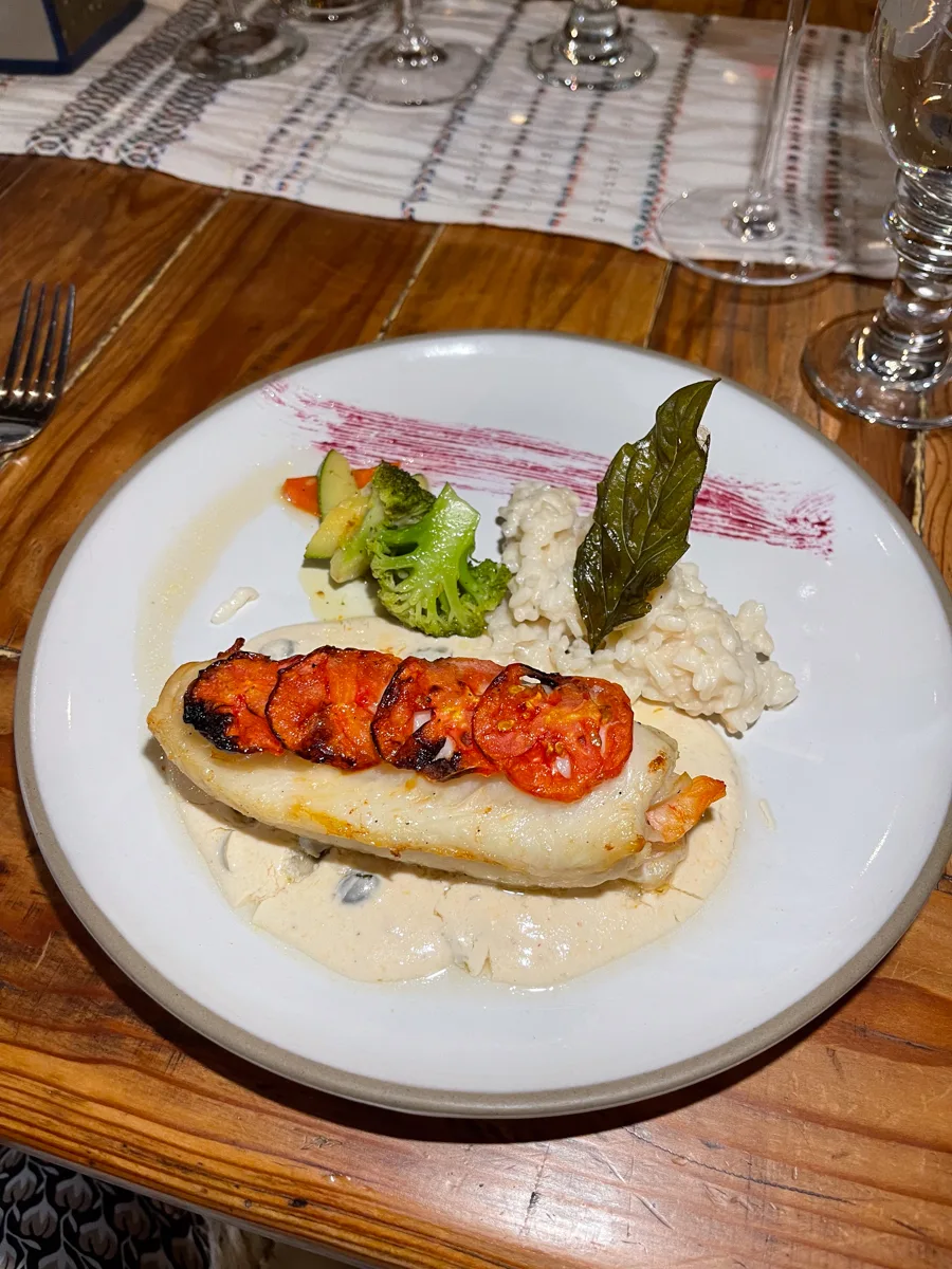 a plate of food with a large piece of fish with slices of tomatoes on top of it that have been roasted. Next to the fish is a small amount of risotto and some vegetables like broccoli.