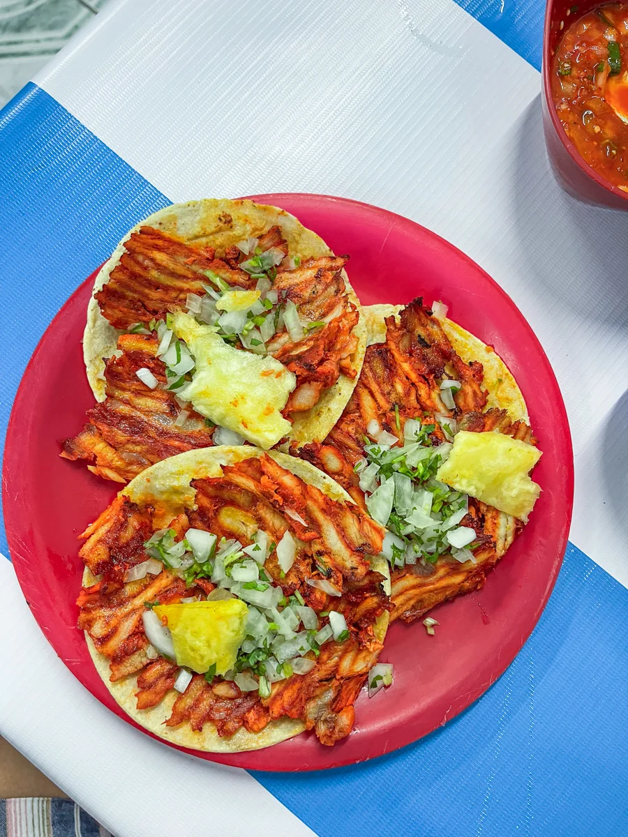 three pastor tacos with a slice of pineapple on top of each one with cilantro and onions on top all on a red plate and the red plate is on a white and blue striped tablecloth