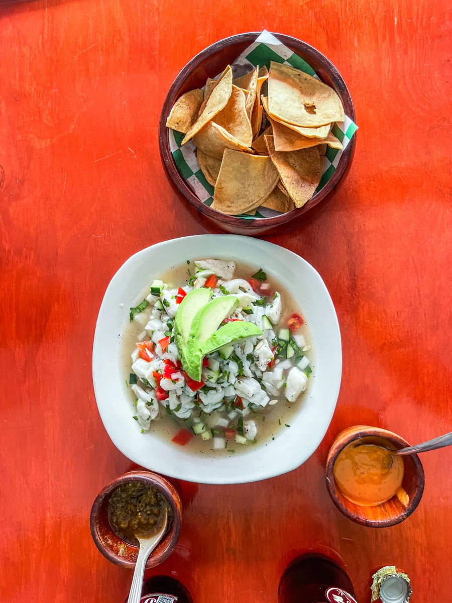 bowl full of fish ceviche with a sliced avocado half on the top and a black bowl on the side filled with tortilla chips. On the other side of the ceviche bowl is two small brown bowls filled with different salsas and each has a spoon in it.