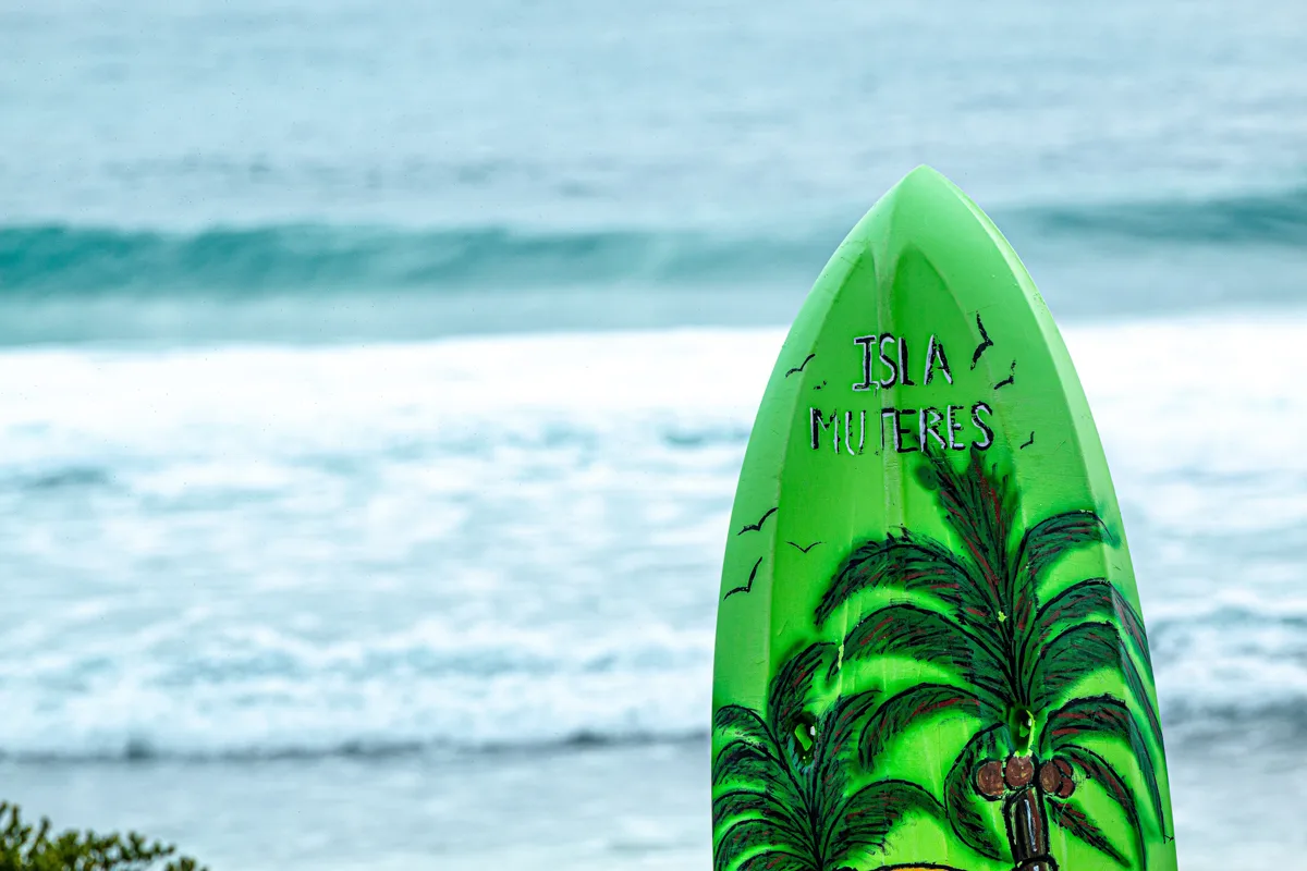 surf board standing in the sand in front of the icea with a palm tree painted on it in one of the best pueblos mágicos in Quintana Roo, Isla Mujeres.