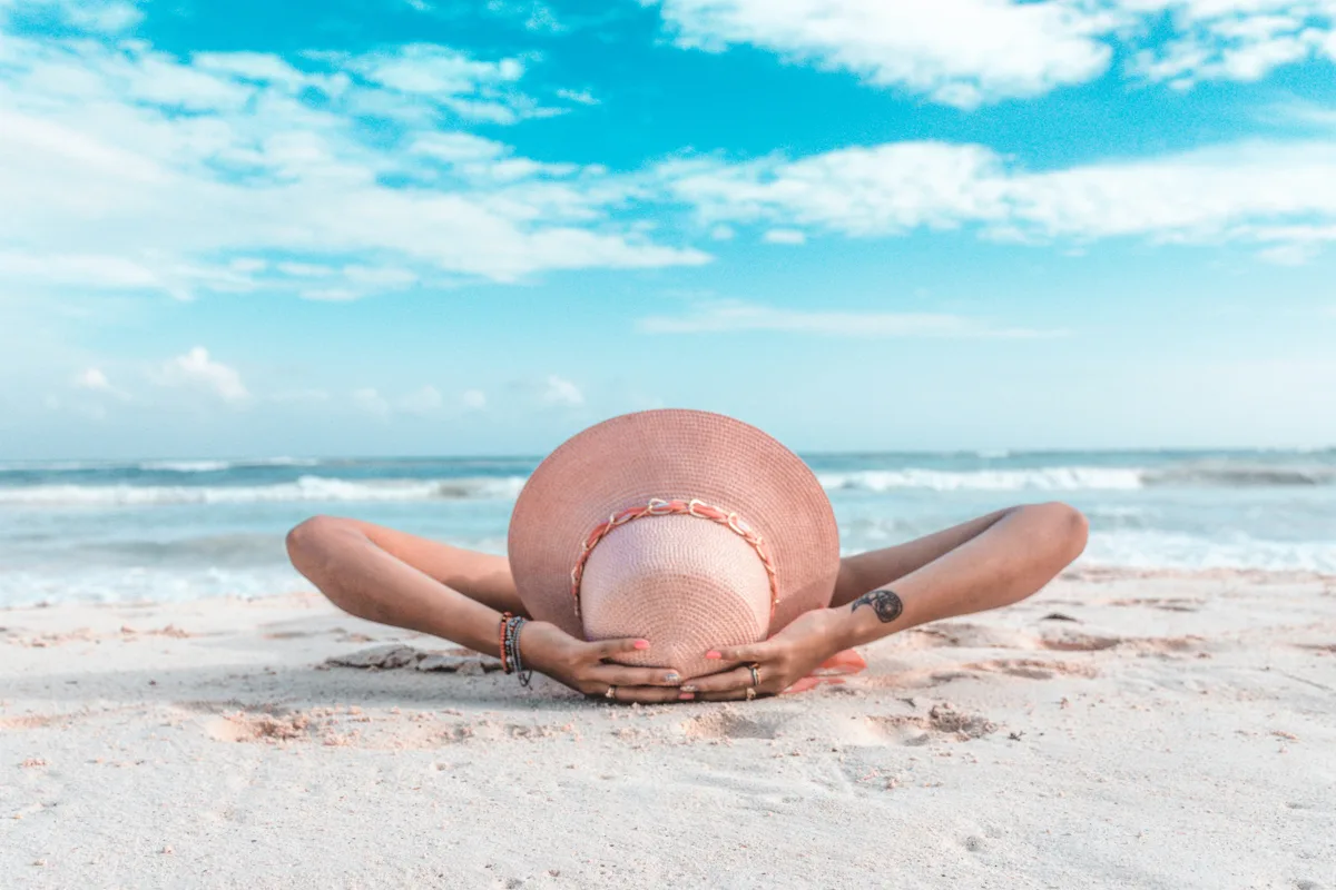 woman laying on the sand with a sunhat on blue skies above her and the ocean near her feet.