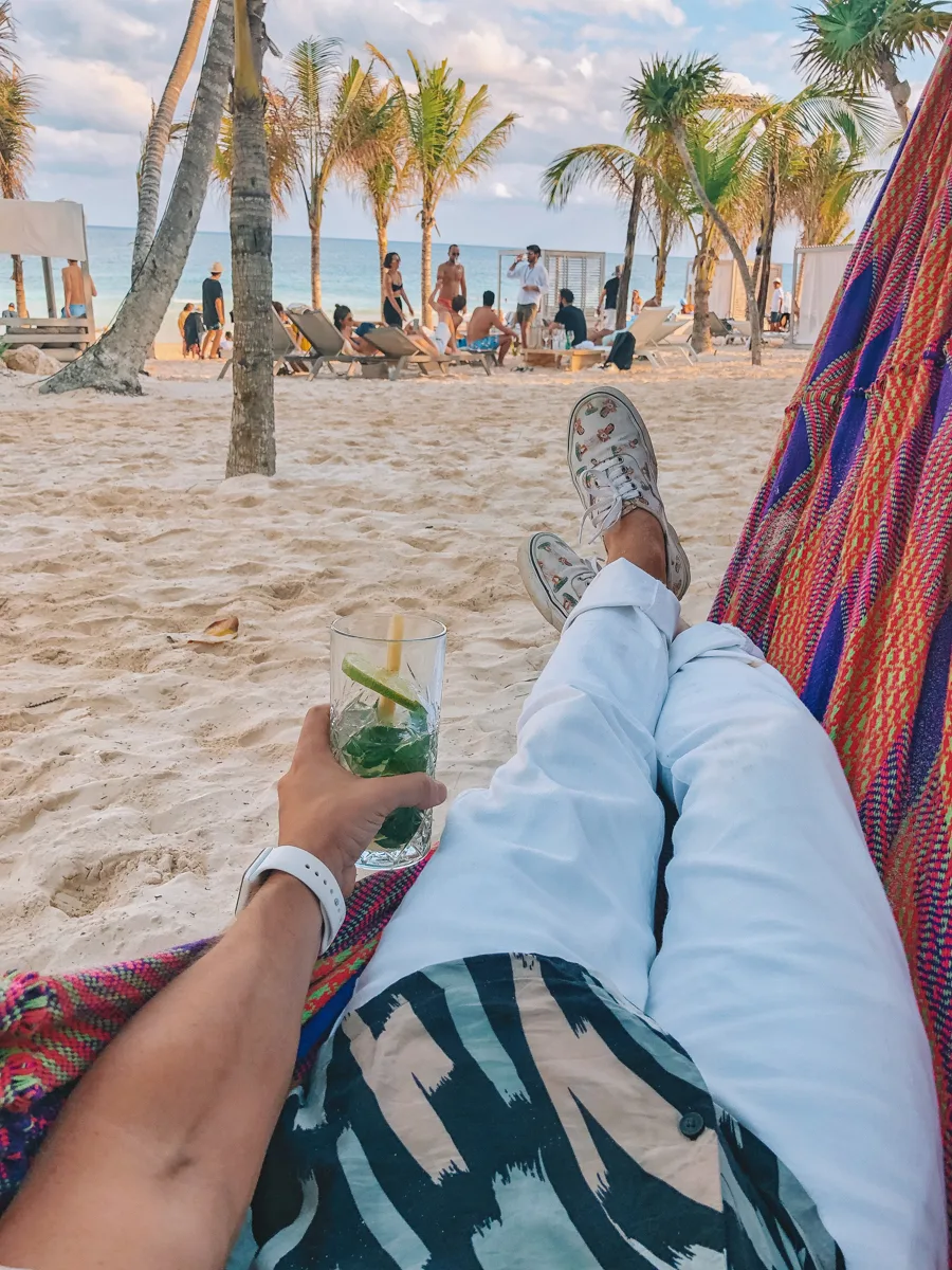 man in a hammock with a drink and the beach in front of him.