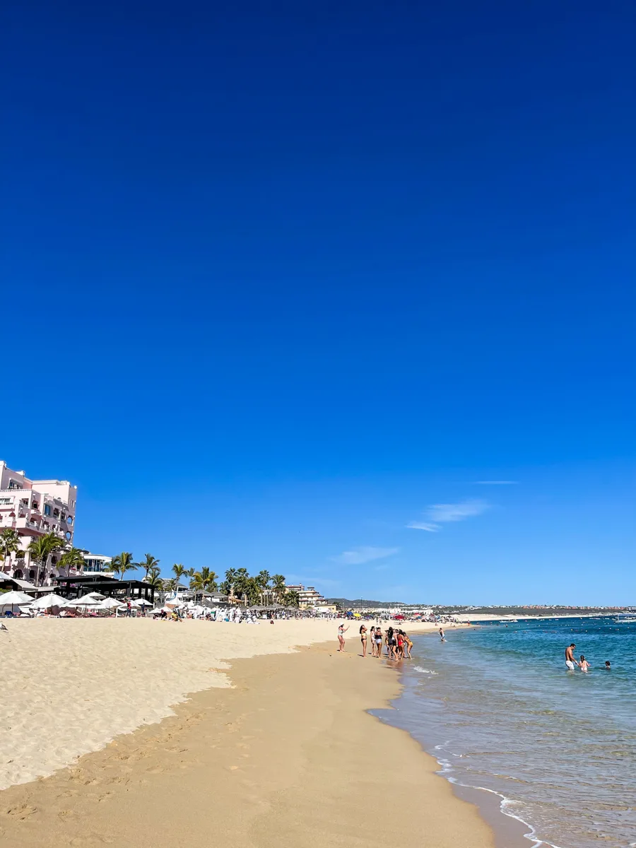 Medano Beach in Cabo San Lucas with bright blue skies and hotels to the left of the sand with bright blue water to the right.