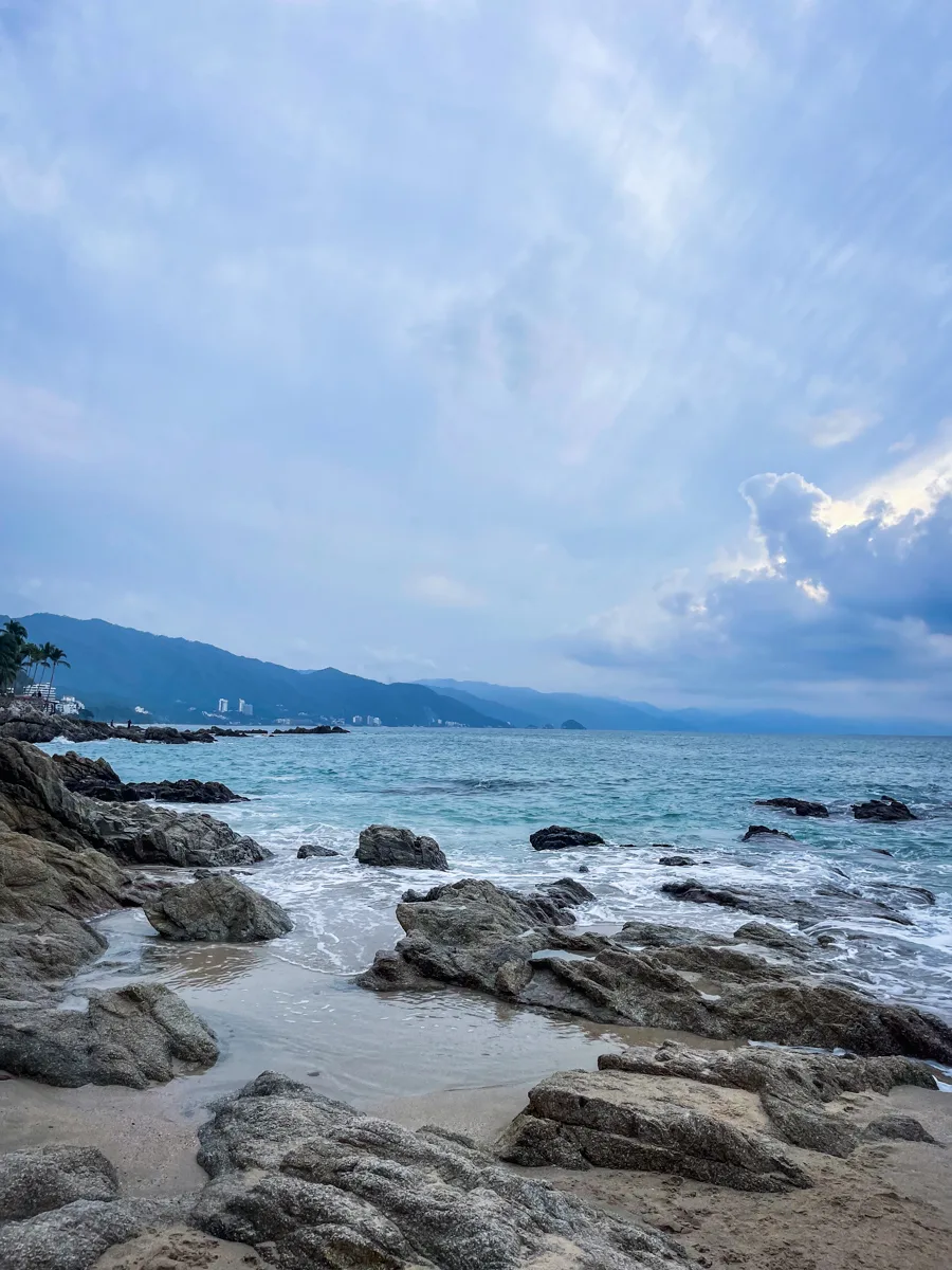beach with some rocks and mountains in the background with clouds above.