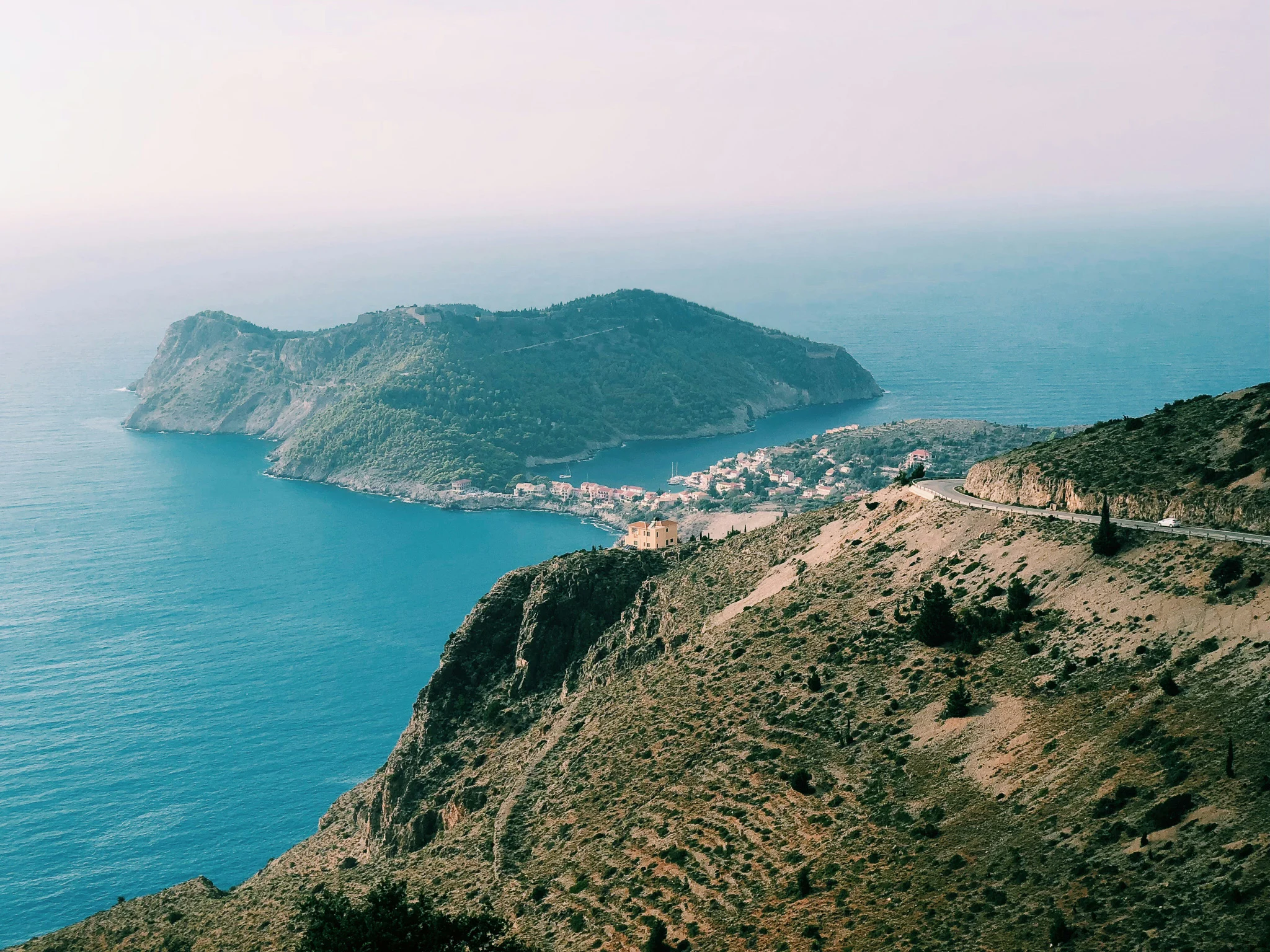 view of an island in the distance with a strip of land connecting it to the land mass in the foreground all covered in trees and surrounded by deep blue water.