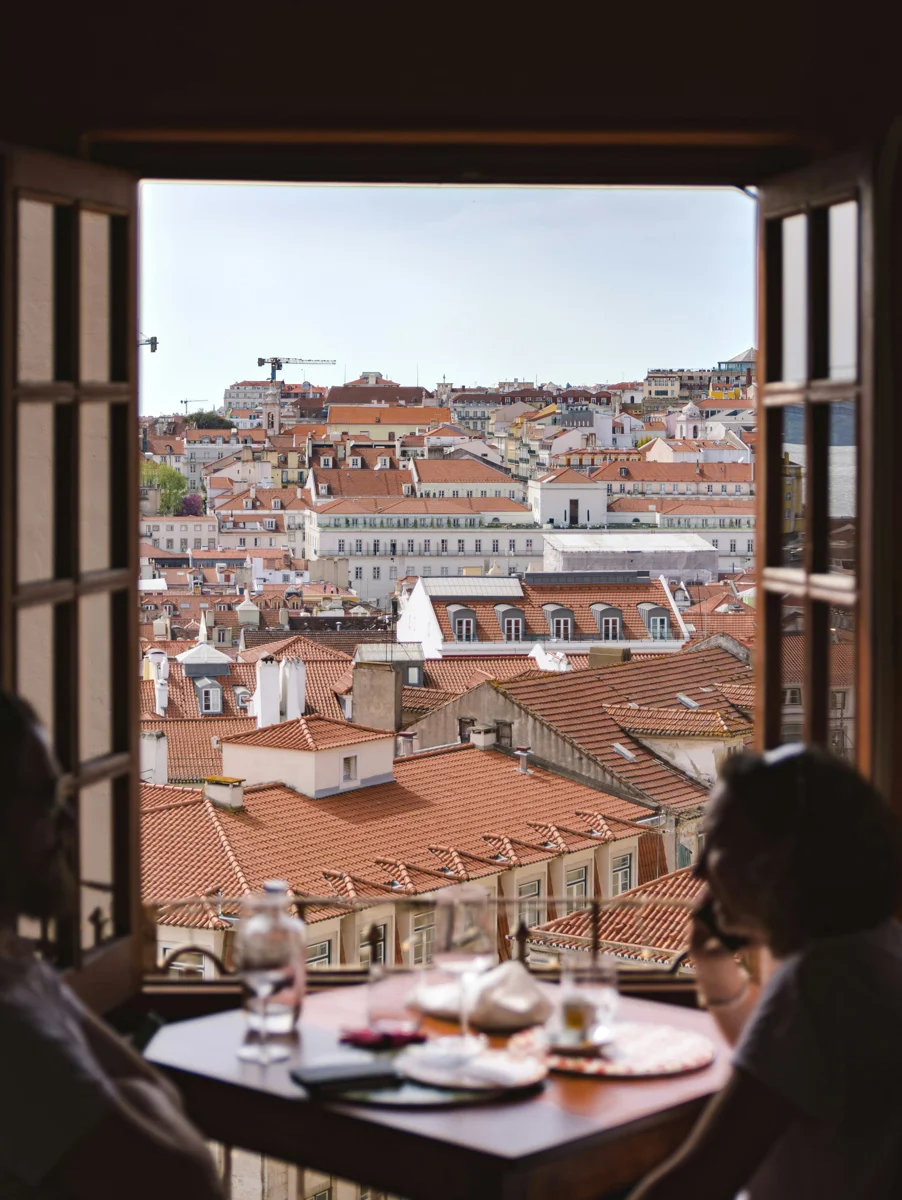 two people sitting at a table in front of an open window with lisbon rooftops and breakfast on the table.