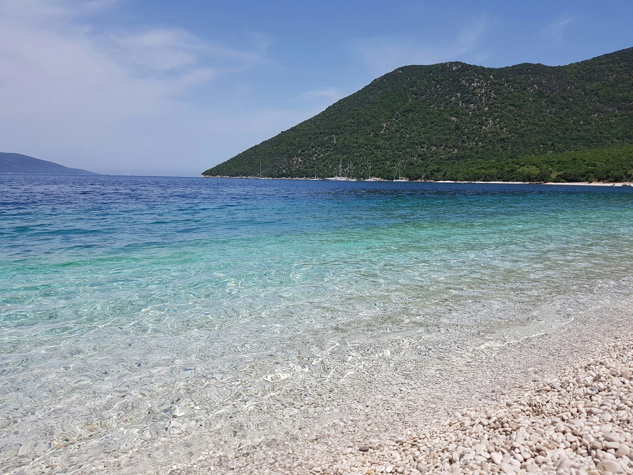 pebbly beach with clear water lapping the shore and changing from clear to turquoise to deep blue as the water gets deeper, a green island in the background all at one of the best beaches in Kefalonia.