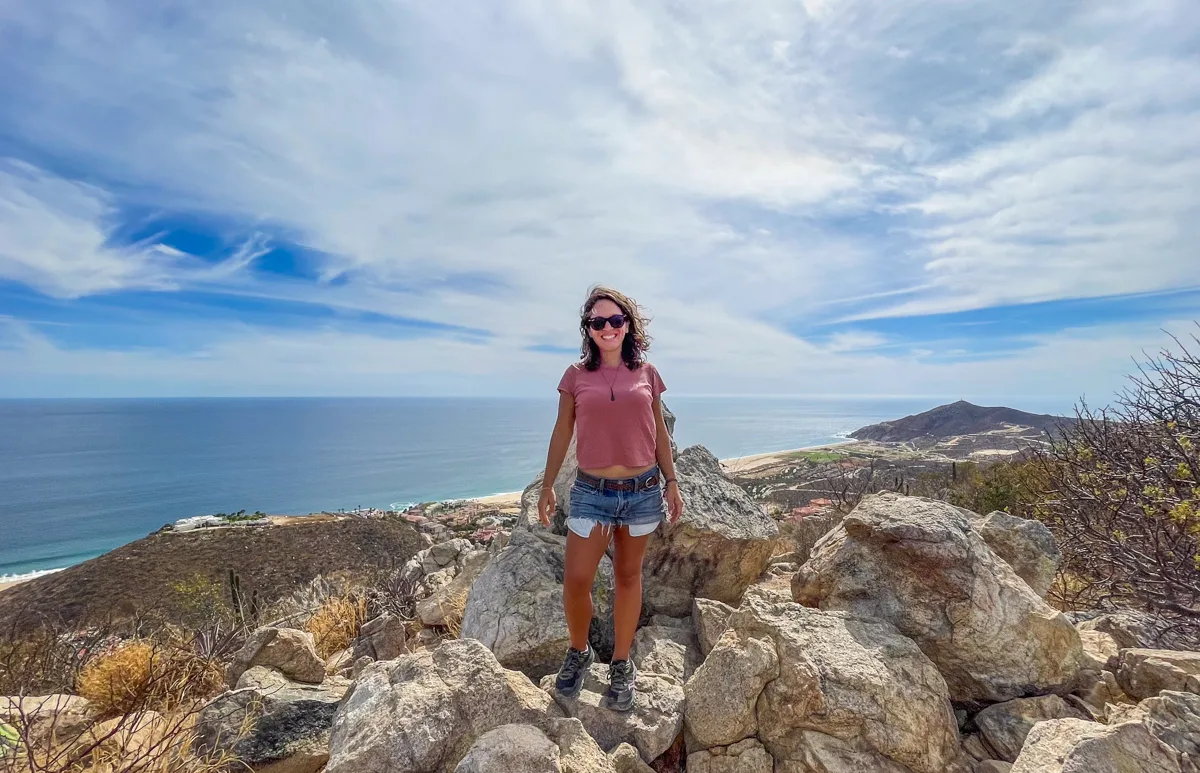 woman standing on a rock on top of a hill with views back over los cabos and the ocean below.