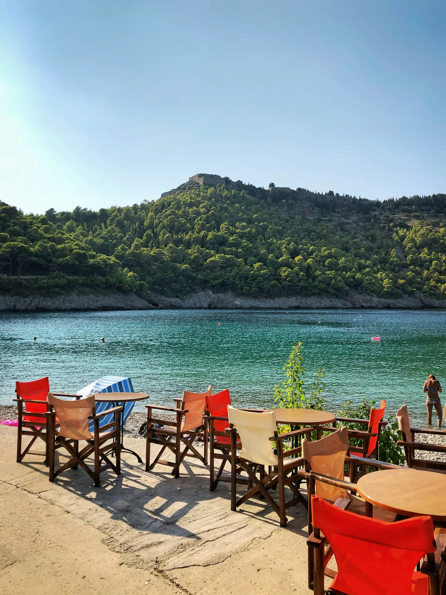tables and chairs on the beach with clear turquoise-green water behind it and a lush green island in the distance at one of the best beaches in Kefalonia.