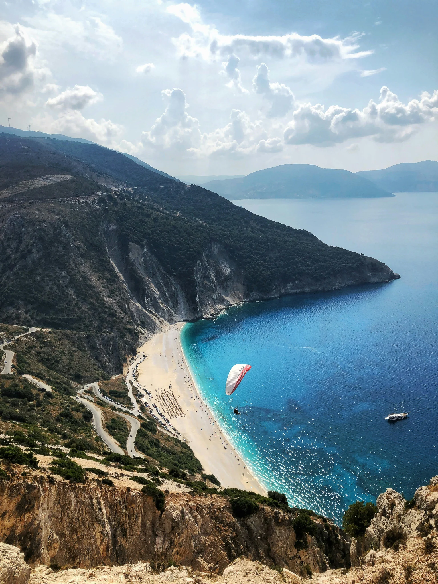 beach at the base of a cliff with a person paragliding over the coast and turquoise water against the shore.