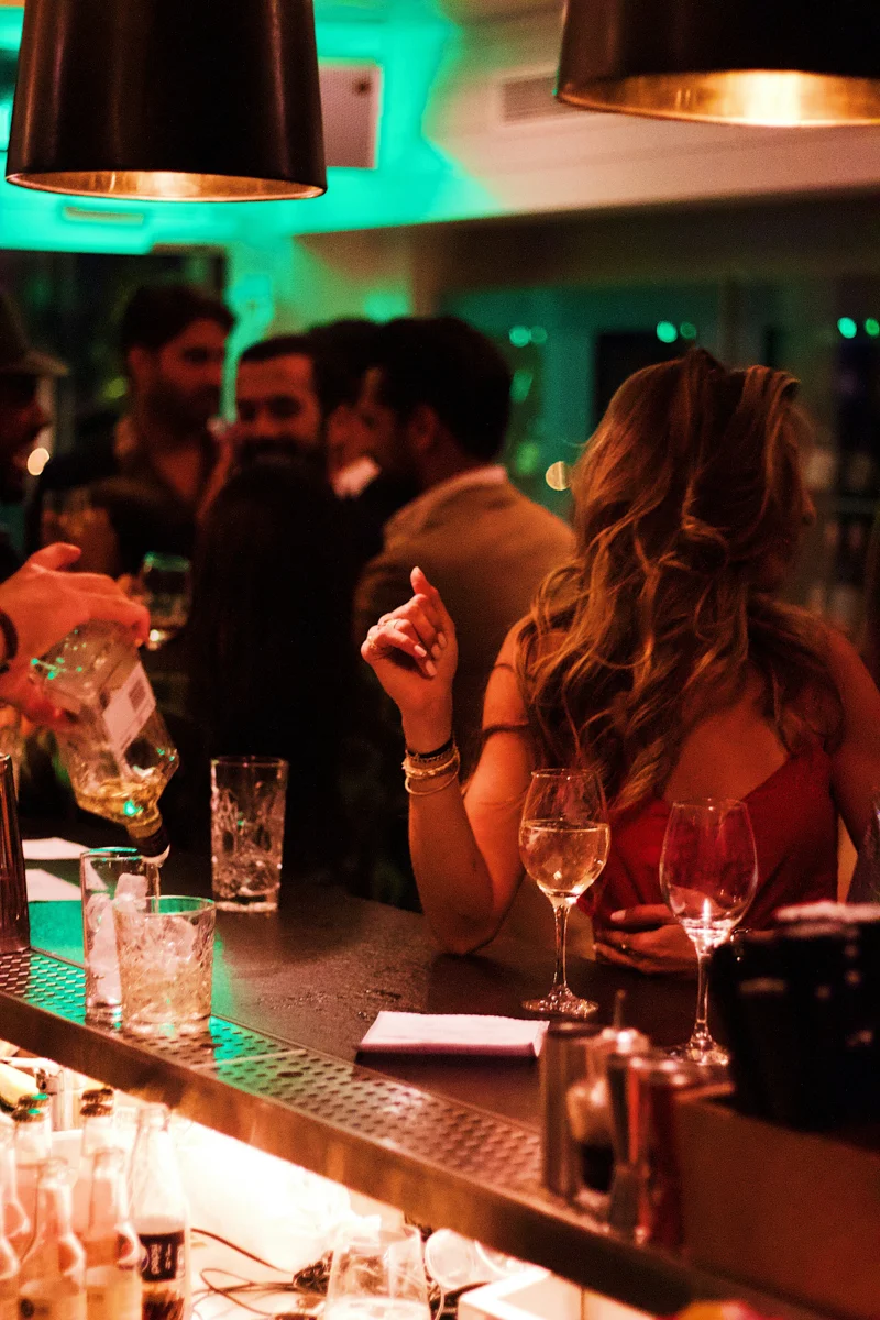 woman standing at a busy car with a crowd of people behind her and a few drinks just visible beneath the back of the bar.