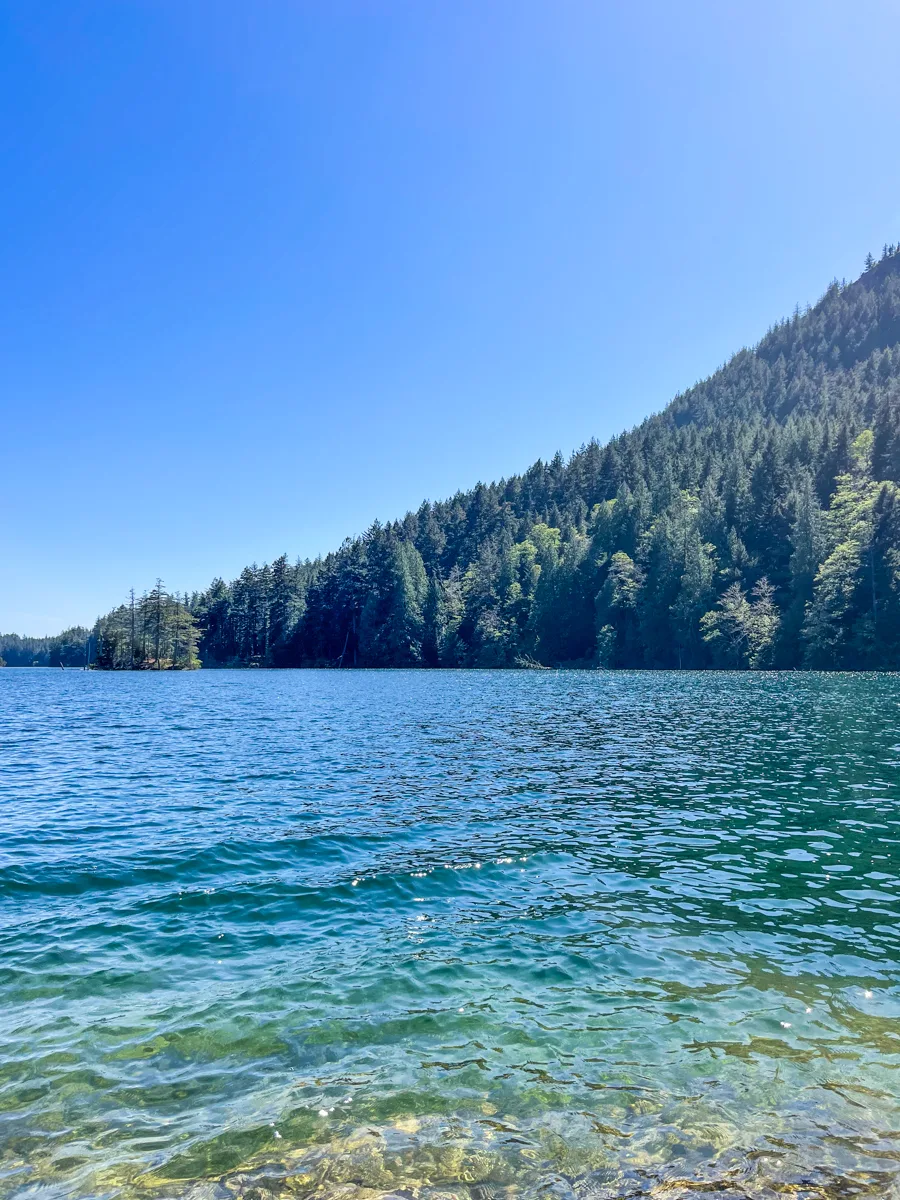 large lake with deep blue water in the middle and shallow light blue edges close to the bottom of the photo with a hill of evergreen trees in the distance.