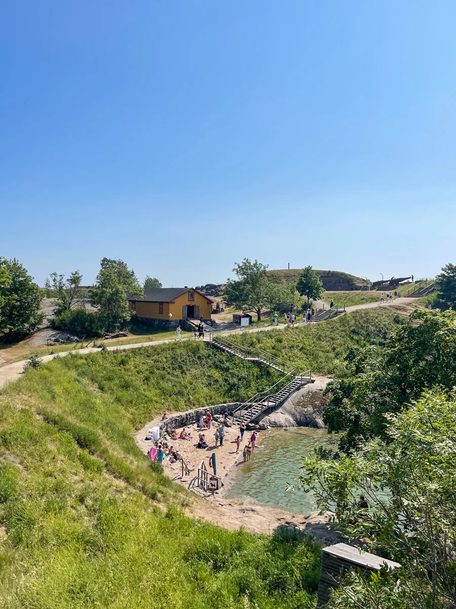 small beach at the base of a cove with a grassy hill above and a winding path along the top of the hill.