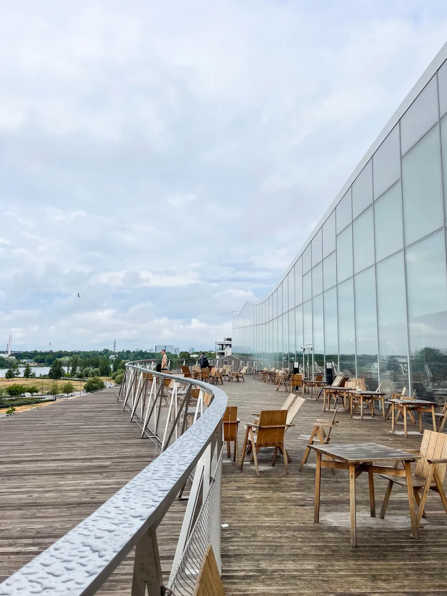 outdoor deck area with a curved railing and wooden chairs and tables on a wooden deck with a glass building to the right.