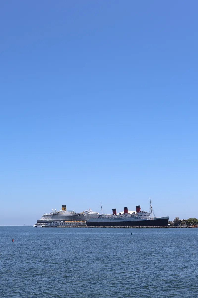 bright blue skies above the horizon and the Pacific ocean below and in the middle of the horizon line are two ships, old and new - an old British oceanliner on the right and a large modern cruise ship on the left.