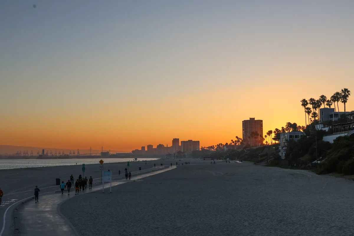 sunset over the beach with palm trees on the right side, the waves calmly crashing to the shore, people walking along the sand and a bright orange line along the horizon.