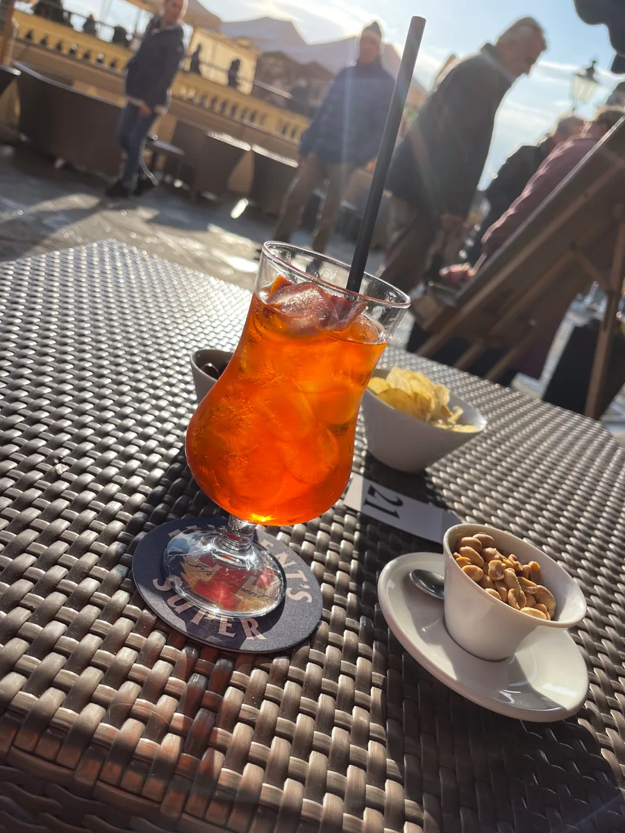 a glass of aperol spritz on a table with a bowl of snacks and the sun shining above.