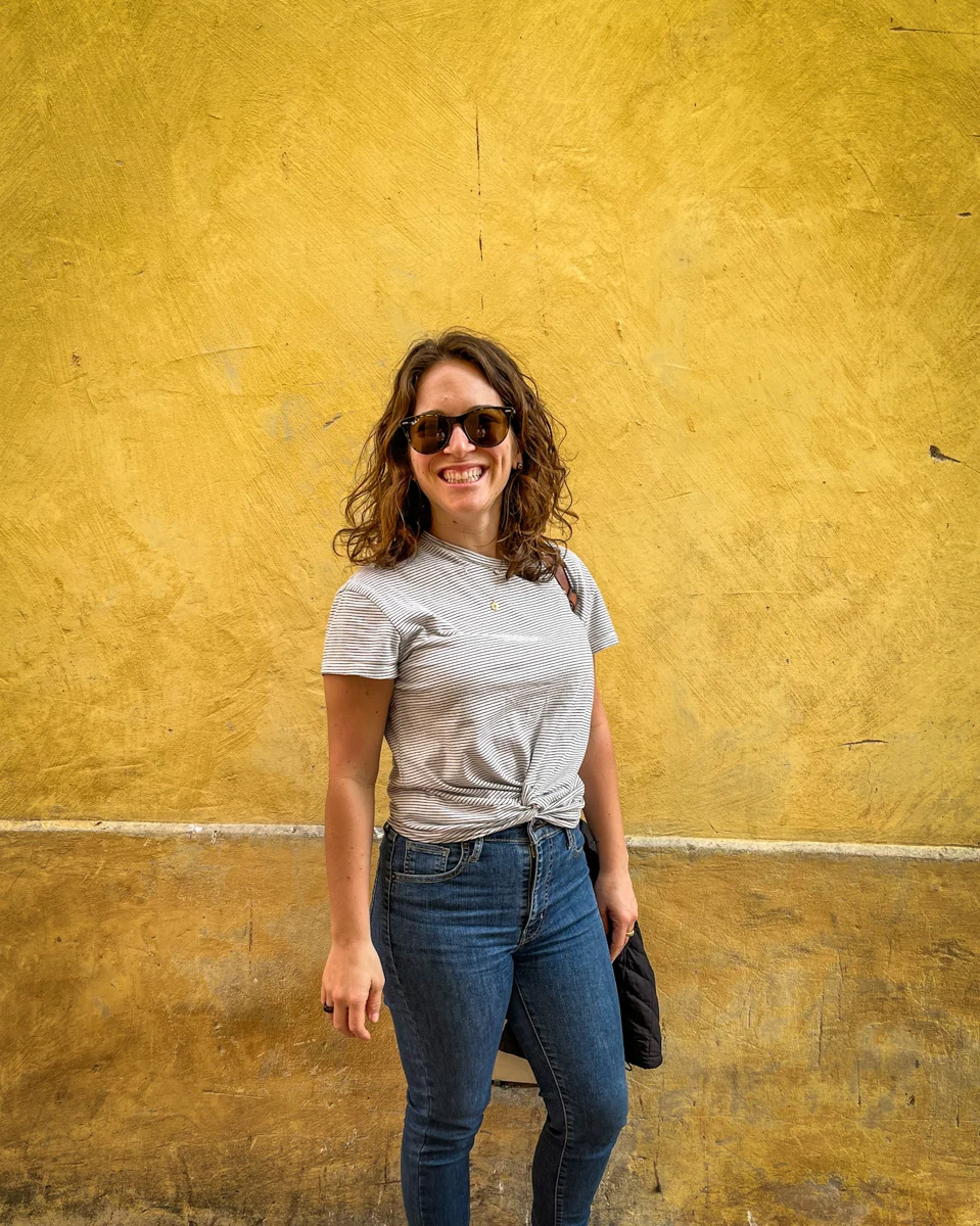 woman in a tshirt and pair of jeans in front of a bright yellow wall.