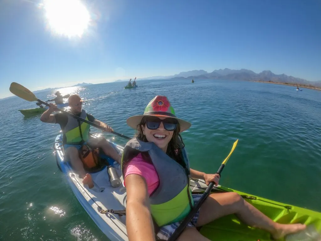 woman sitting in a kayak with a pink tshirt on, a life jacket and a big brimmed hat with a flower painted on the front.