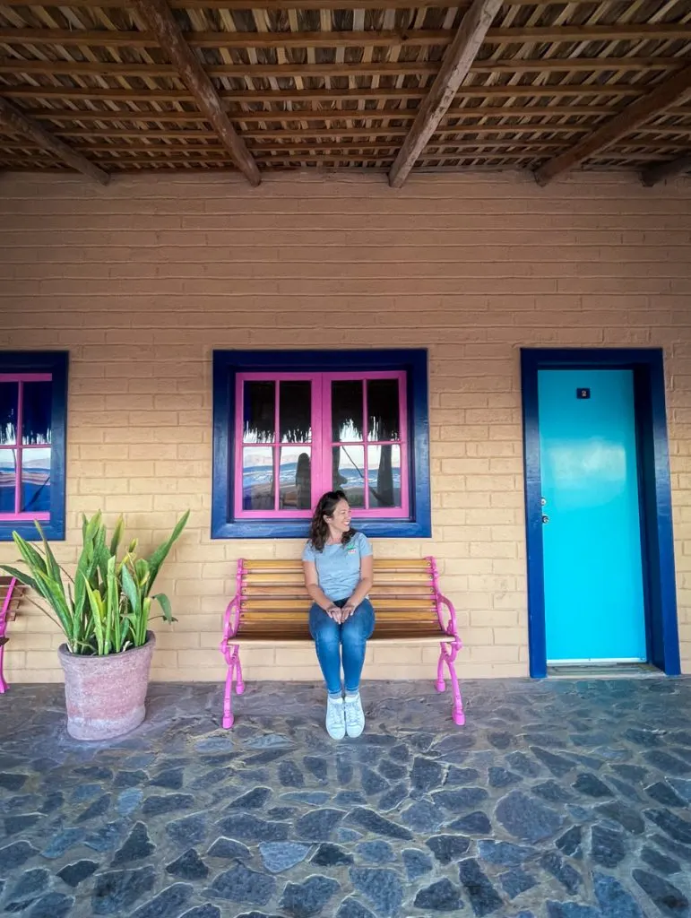 woman sitting on a bench in front of a colorful building with a bright blue door to her right and a bright green plant to her left.