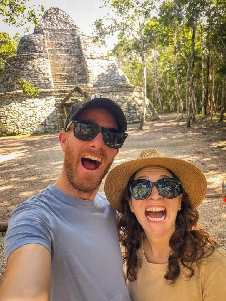 man and woman taking a selfie together in front of a stone pyramid in Mayan style behind them. They have their mouth's open like they are about to shout in excitement.