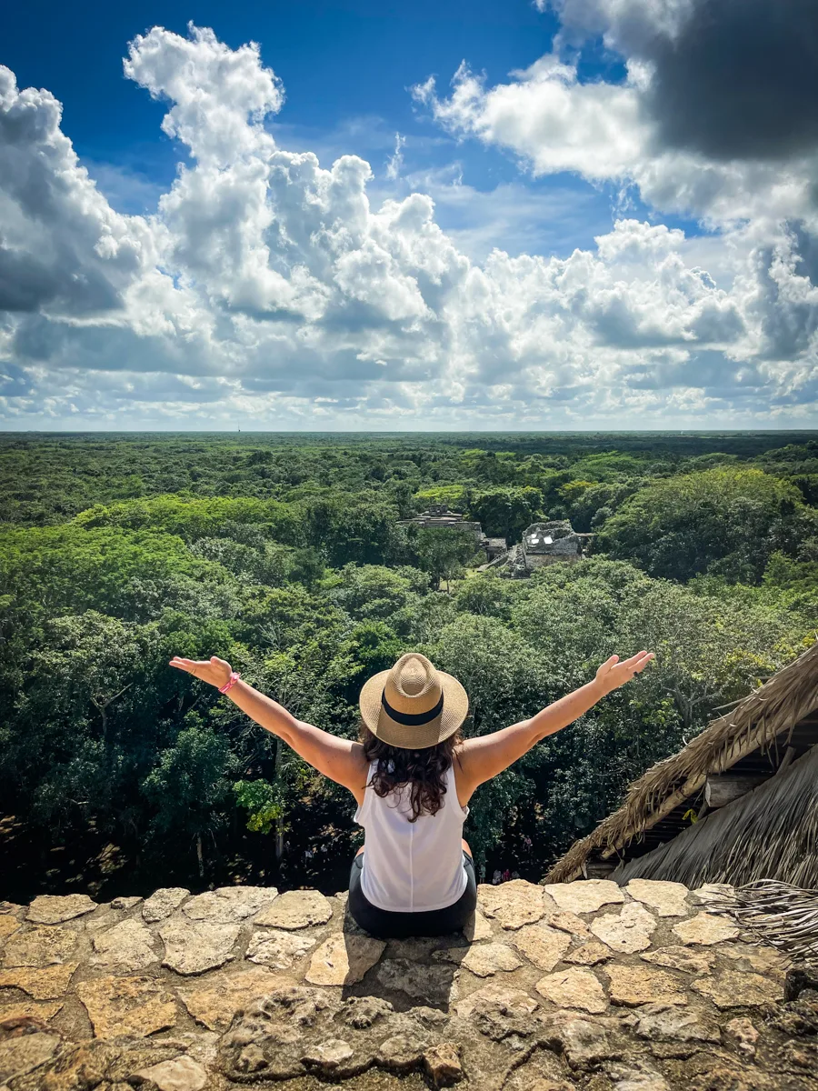 woman sitting on top of a stone step with a flat jungle in front of her, but you can only see the tree tops from behind her.