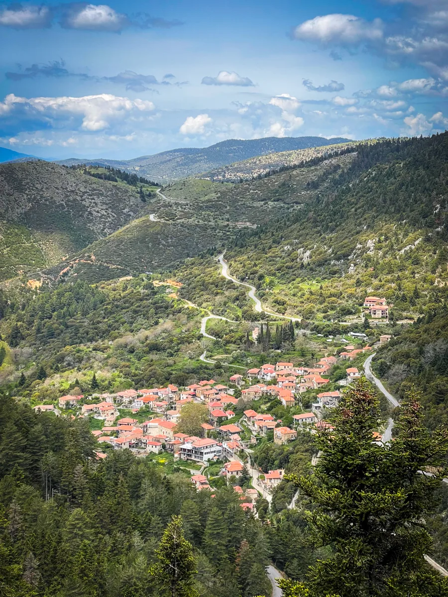 views of a small greek town from above in the mountains. There are red roofed buildings below tucked into the valley surrounded by lush green hills and the occasional road that cuts through the landscape.