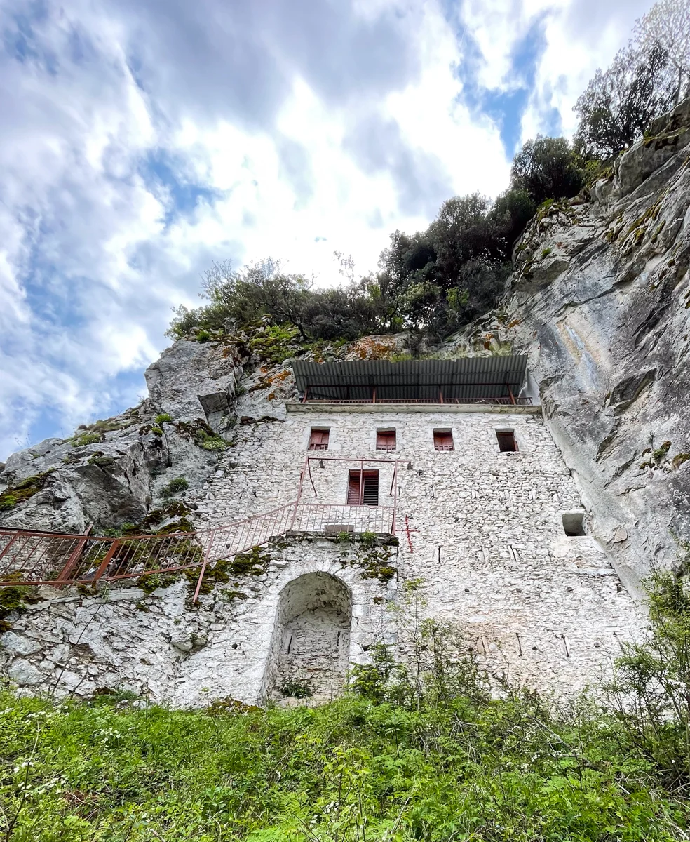 a church carved into the stone of the side of a cliff, all white, with four small windows in a row at the top under a small metal roof and a metal staircase leading to the small red door on the left.