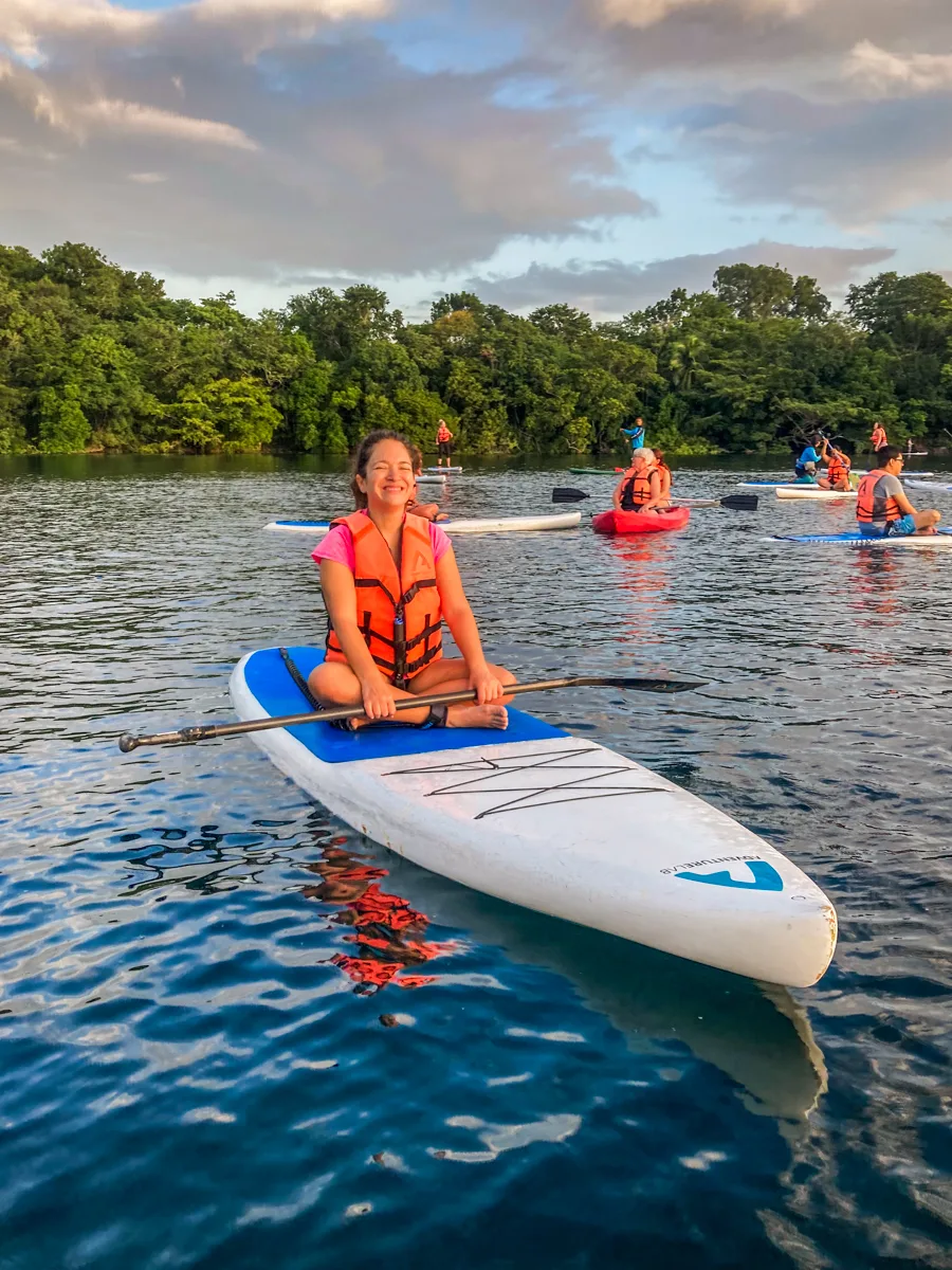 woman sitting on a paddle board in the ocean with a row of trees on the horizon behind her.