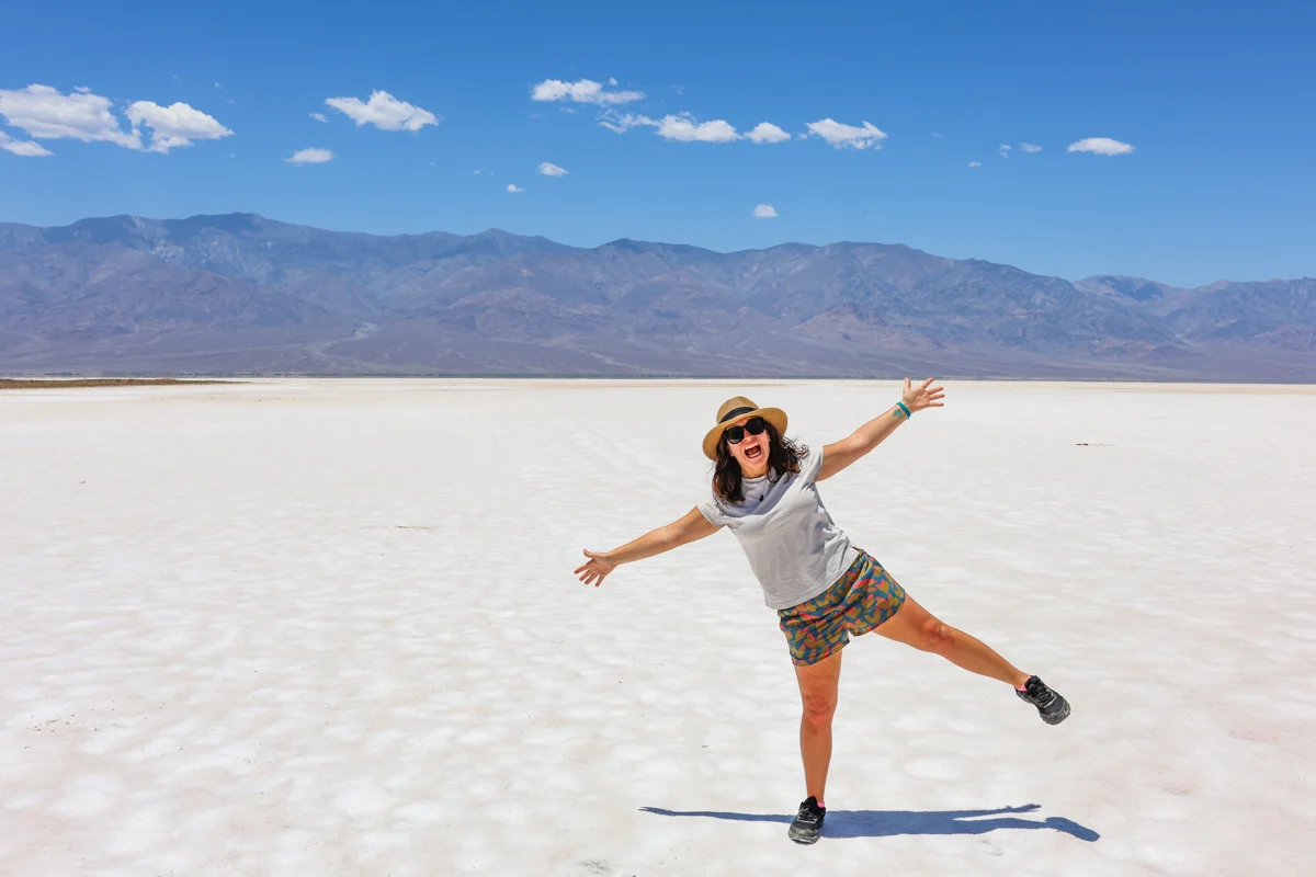 death valley with a woman standing in the middle of badwater basin with mountains in the background and bright white salt flats on the ground.