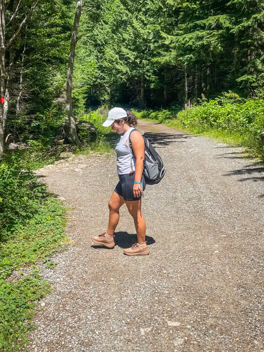 woman walking on a trail with green forest on each side. she has a hiking backpack and shorts and a tanktop on.