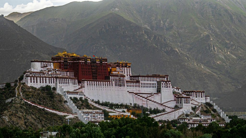 palace in the mountains surrounded by huge green peaks all around it. the palace is white except for the middle part which is a deep red color like the color of a monk's robes.