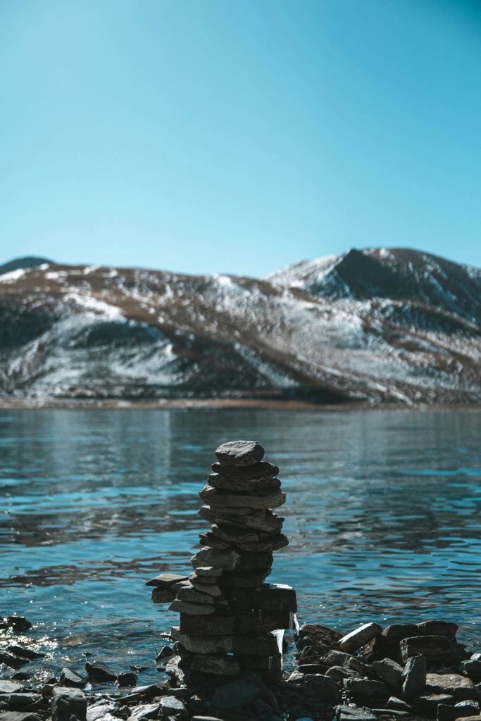glacier lake in tibet with a stack of rocks in the foreground and the clear turquoise waters and mountain ranges in the background.