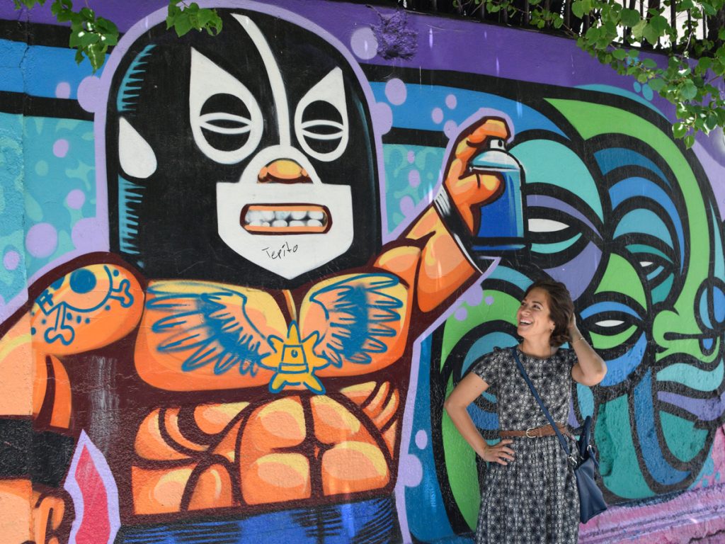 woman standing in front of a spray painted wall with the image of a Lucha Libre fighter wearing a mask and showing off his muscles while spray painting the same wall.