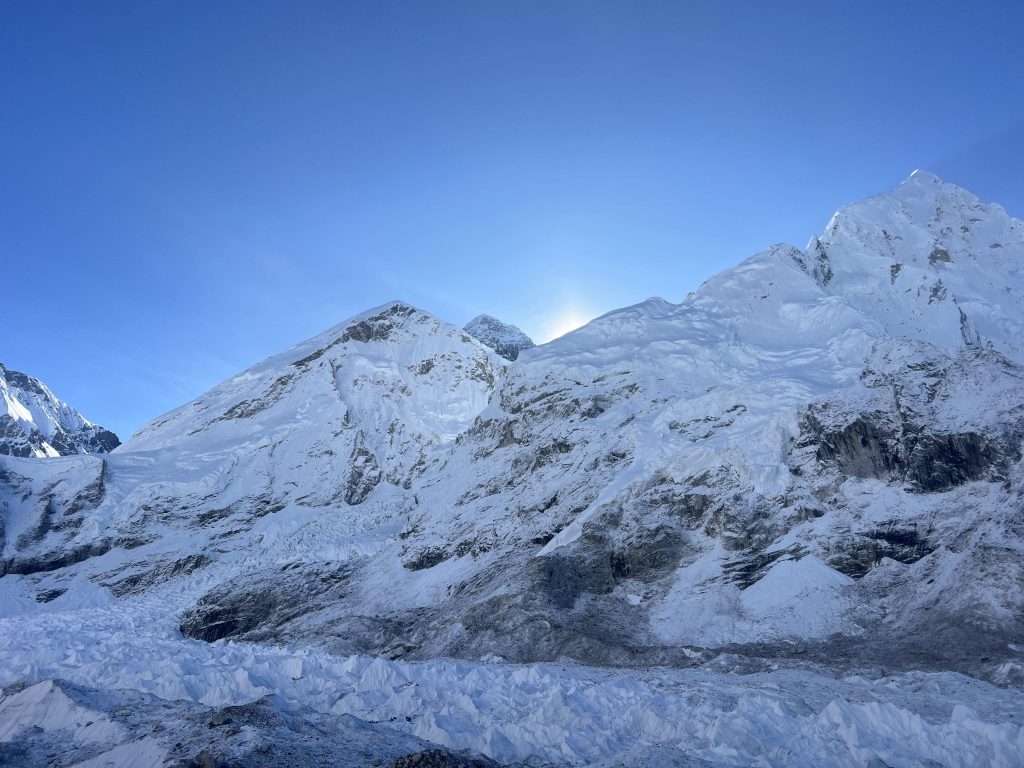 everest base camp in nepal with the sun hiding behind snow covered mountains and bright blue skies above.